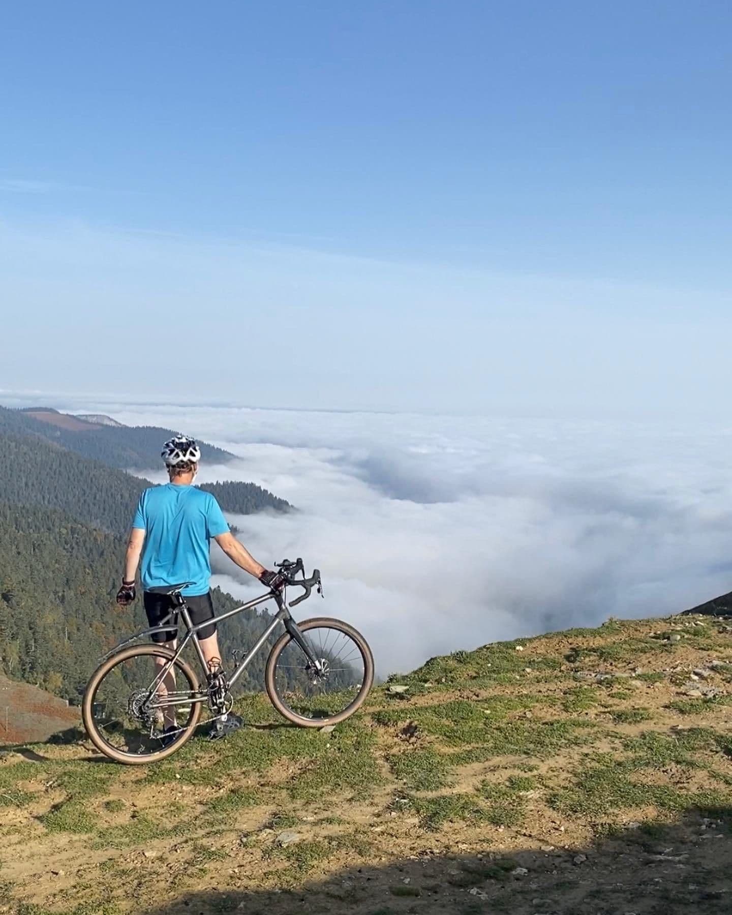 A cyclist stands beside a gravel bike on a grassy ridge, looking out over a sea of cloud filling the valleys below.
