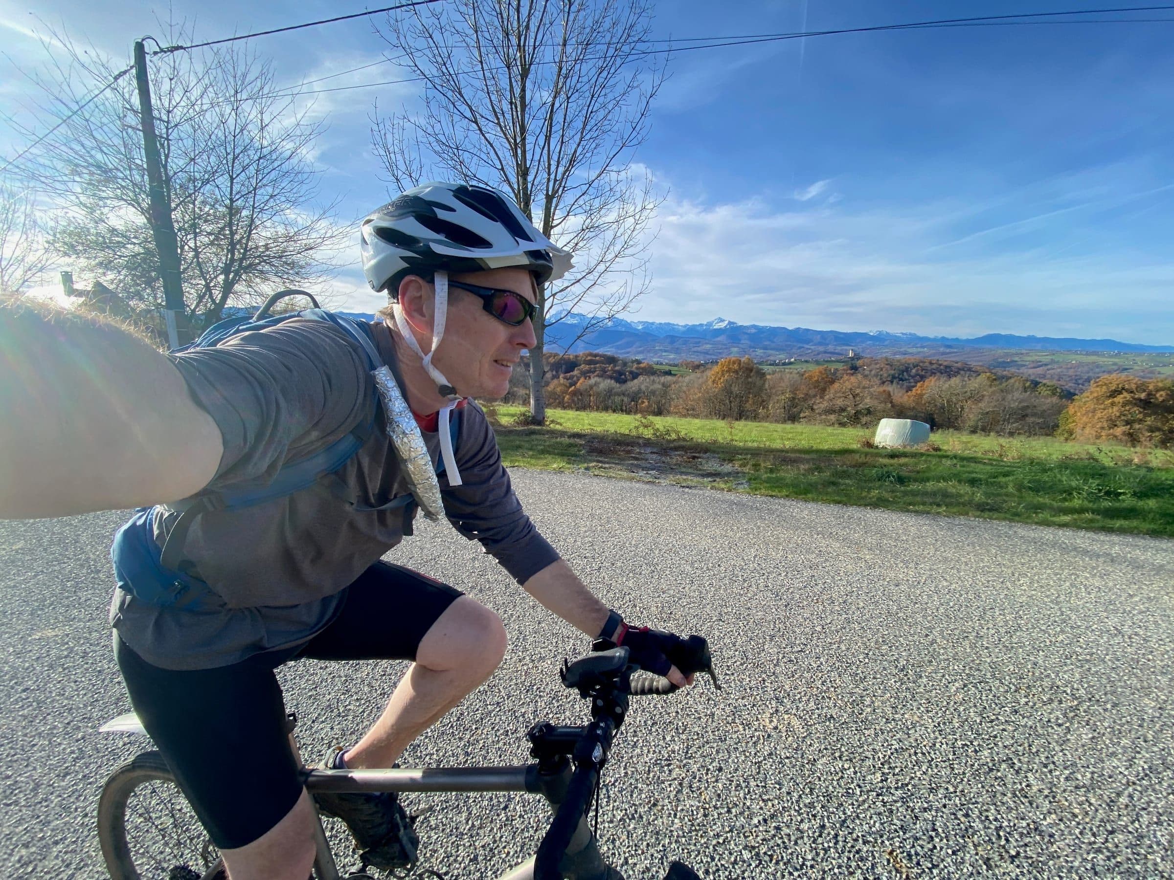 A cyclist takes a selfie on a country road, with autumn trees and snow-capped mountains behind him.
