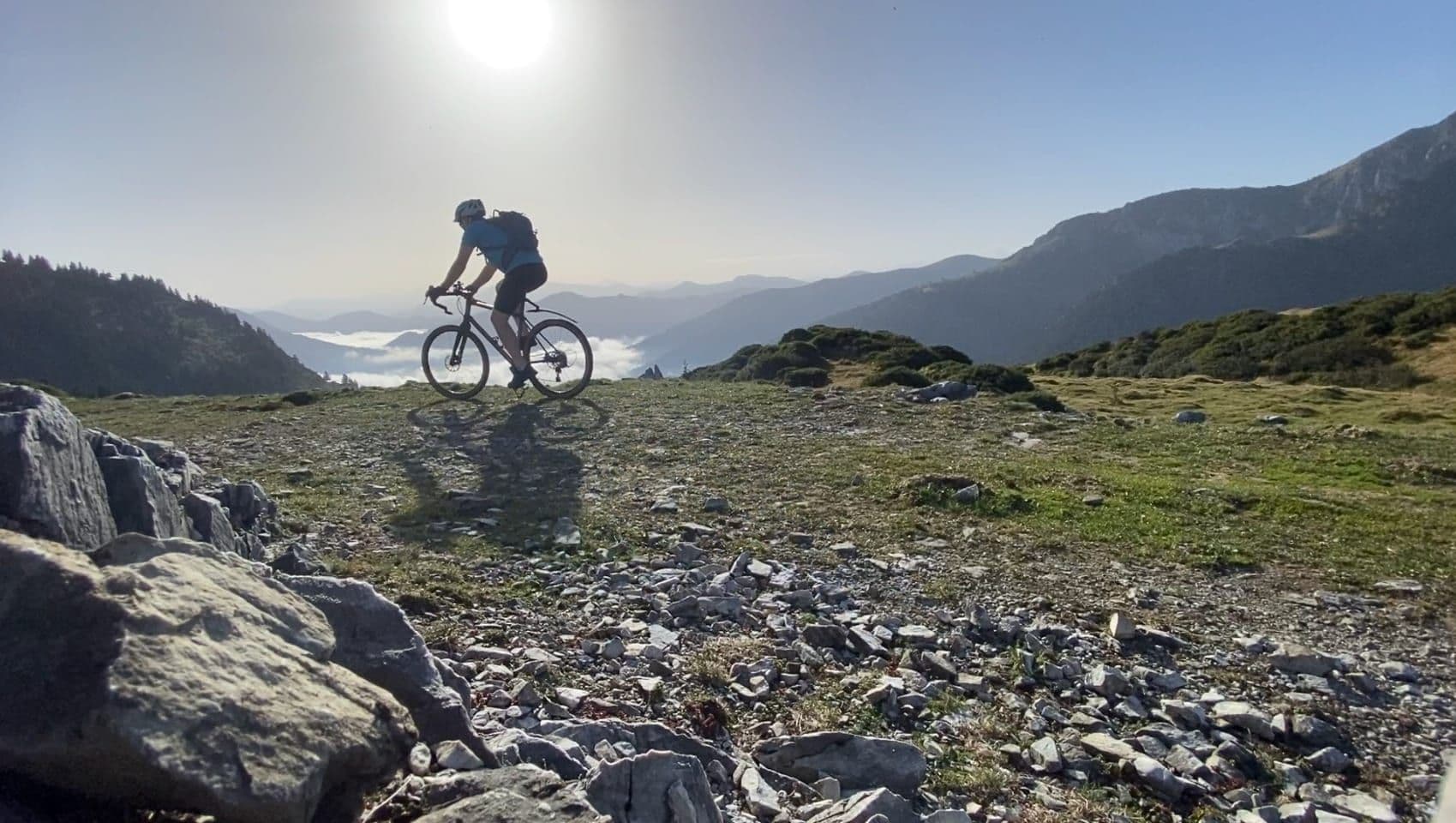 A cyclist silhouetted against the morning sun crosses a rocky high pasture, with cloud lying in the valleys beyond.