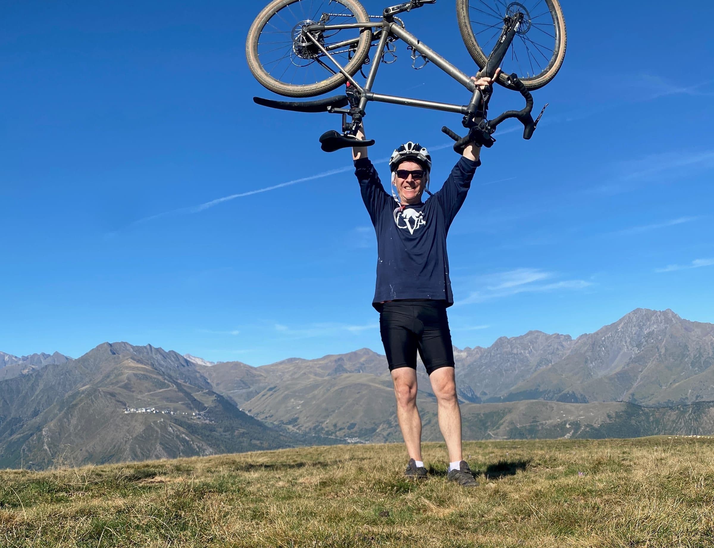 A smiling cyclist holds his bike high over his head on a grassy summit, with mountain peaks behind him.